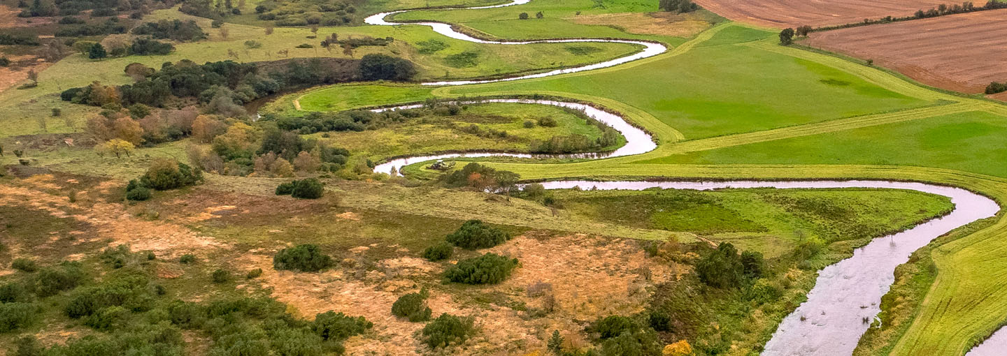 Luftfoto af et snoet vandløb gennem et varieret landskab med marker, græsområder og små skovarealer, der illustrerer balancen mellem landbrug og natur i det danske landskab