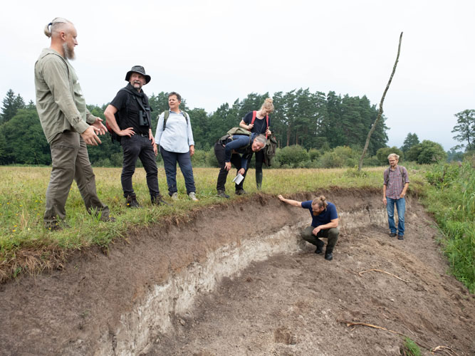 En gruppe mennesker står ved kanten af en dyb udgravning i landskabet. En person undersøger jordlagene, mens de andre betragter og diskuterer den synlige jordprofil.