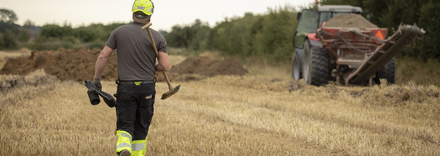 Mand i arbejdstøj går hen over en stubmark i retning af en større opgravning, hvor en traktor holder og venter