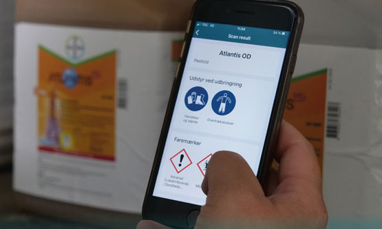 Nærbillede af en mobiltelefon i en hånd. på mobilskærmen ses grafiske og ikoner og advarselsikoner. I baggrunden anes store plastikdunke med noget indhold.